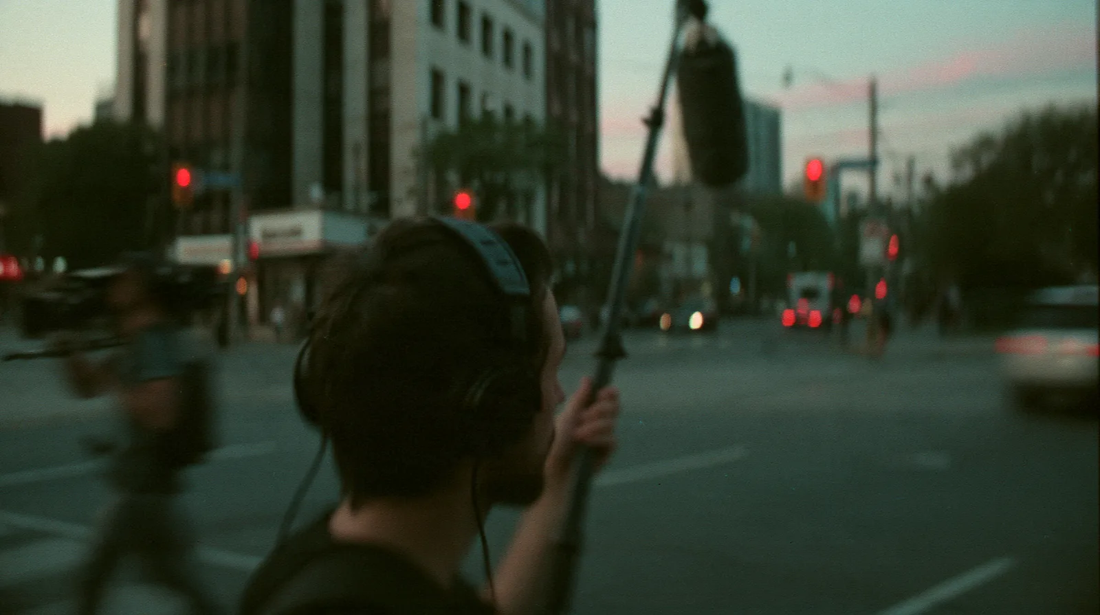 A sound recordist in headphones holding a boom microphone pole on a city street at dusk